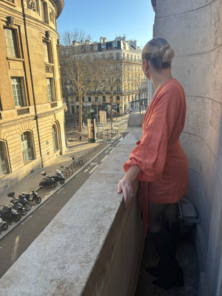 Woman in an orange dress standing on a balcony overlooking a city street with buildings and parked motorcycles.